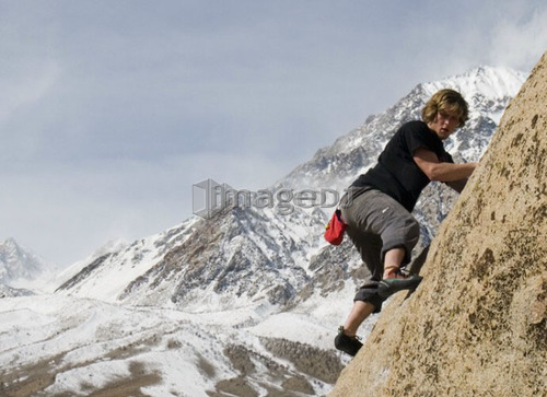 Man (20-25) climbing a rock face with a snow-capped mountain in the background, Bishop, California