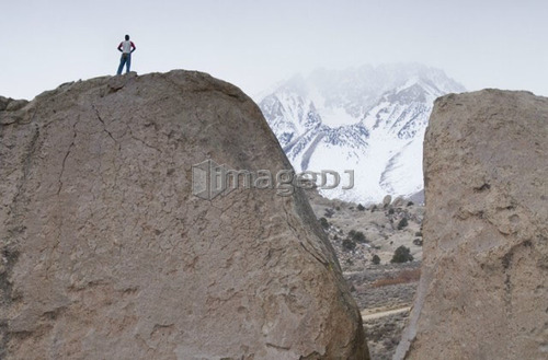 Man (20-25) standing on top of a large boulder with a snow-capped mountain in the distance partially hidden by fog. Bishop, Cali