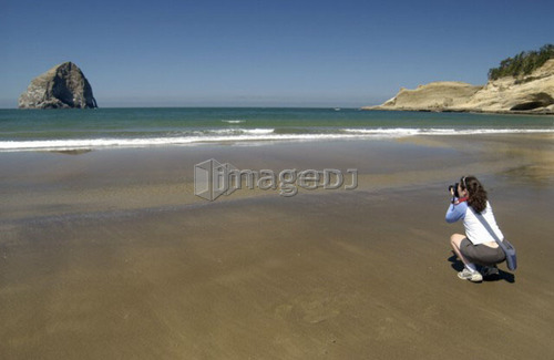 Woman (20-25) photgraphing on a sandy beach with blue sky and rock formations, Newport, Oregon