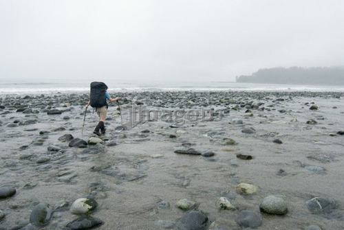 Woman (20-25) hopping across a creek with a large backpack on with mist in the background, West Coast Trail, Pacific Rim Nationa