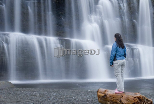 Woman (20-25) standing on a beach log in front of a waterfall