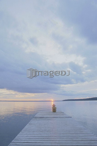 Woman sitting on dock at sun set Clear Lake; Canada, Manitoba, Riding Mountain National Park