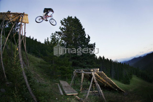 Mountain biker jumping from wooden ramp on to log ramp; Canada, Alberta, Moose Mountain