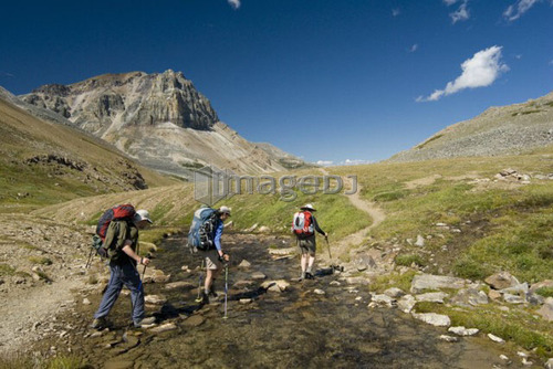 Three hikers cross a small creek in an alpine meadow.; Canada, Alberta, Jasper National Park, Skyline Trail
