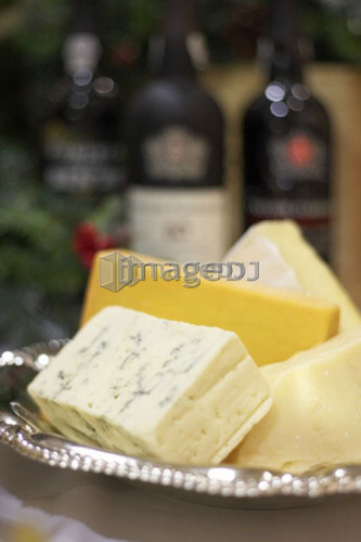 Bricks of blue, cheddar, parmesan, and brie cheese in a silver dish with bottles of port in background