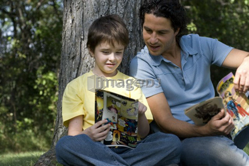 Father and son beside tree reading comic books, Assiniboine Park, Winnipeg, Canada