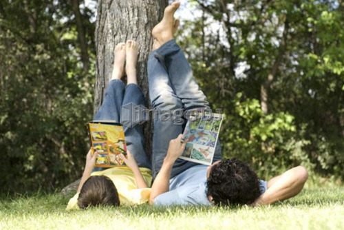 Father and son lying on backs with feet resting on tree, reading comic books, Assiniboine Park, Winnipeg, Canada