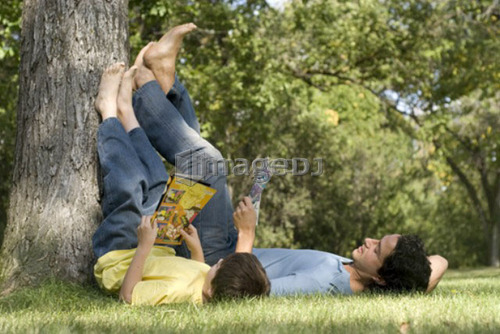 Father and son lying on backs with feet resting on tree, reading comic books, Assiniboine Park, Winnipeg, Canada